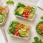 Vier verschiedene gesunde Lunch-Boxen mit frischem Gemüse und Vollwertkost auf einem hellen Tisch neben einem Laptop – Meal Prep für die Arbeitswoche.