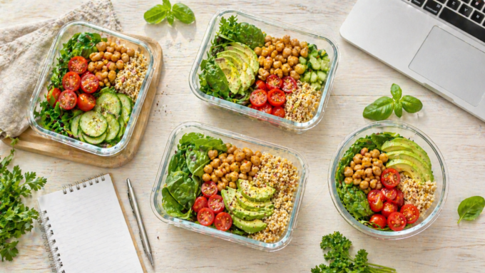 Vier verschiedene gesunde Lunch-Boxen mit frischem Gemüse und Vollwertkost auf einem hellen Tisch neben einem Laptop – Meal Prep für die Arbeitswoche.